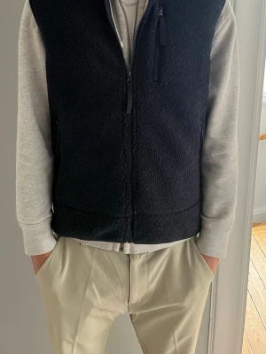 Svart fleeceväst med dragkedja - Svart väst i mjuk fleece med hel dragkedja framtill och hög krage. Västen har en enkel, stilren design och en bröstficka med dragkedja. Perfekt att slänga över en hoodie för en avslappnad look. Stl L men sitter som M 