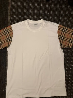 Burberry tröja - Vit t-shirt från Burberry med klassiska beige och svart-rutiga korta ärmar. Tidlös design med rund halsringning och avslappnad passform. Perfekt för dig som gillar stilrena plagg med en twist.