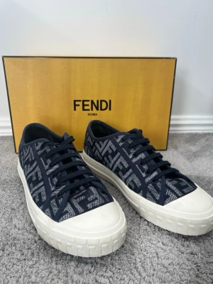Fendi sneakers i blått och vitt - Snygga sneakers från Fendi med blå textil och ikoniskt FF-mönster i vitt. Skorna har vita, robusta gummisulor och matchande vita tåkappor. Klassisk låg modell med blå skosnören och exklusiv känsla. Perfekta för dig som vill sticka ut med designerstil.
