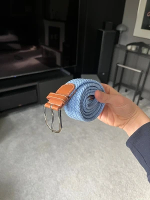 Ljusblått flätat bälte med skinn - Snyggt ljusblått flätat bälte med detaljer i brunt skinn och silverspänne. Perfekt accessoar för att lyfta din outfit med en fräsch färg och stilren design.
