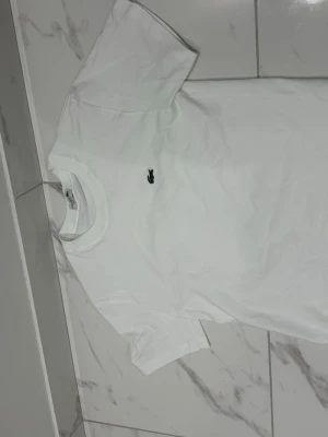 Vit t-shirt från Lacoste - Klassisk vit t-shirt från Lacoste med den ikoniska lilla krokodilloggan broderad på bröstet. T-shirten har rund halsringning och korta ärmar. Perfekt basplagg i mjuk bomull som passar till allt.