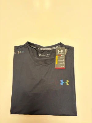  Under Armour t-shirt - Snygg mörkblå t-shirt från Under Armour med korta ärmar och rund hals. Tillverkad i ett lätt och funktionellt polyestermaterial, perfekt för träning. Logga på bröstet och texten 'Under Armour' i regnbågsfärger längs sidan ger en sportig vibe.