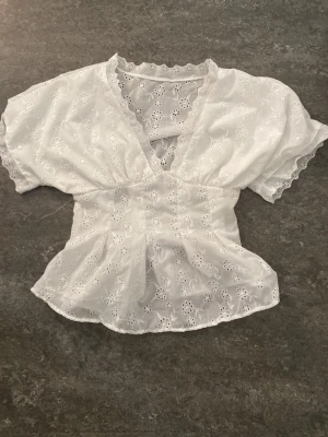 Vit broderad blus med v-ringning - Söt vit blus med broderade blommor och v-ringning. Modellen har korta ärmar, lätt volang nertill och spetsdetaljer längs kanterna. Perfekt för en fräsch och somrig look.