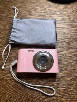 Rosa digitalkamera 2.7K med väska - Snygg rosa digitalkamera med 2.7K video, 40 MP och autofokus. Kameran har en stor display på baksidan, flera knappar för enkel navigering och en praktisk handledsrem. Medföljer gör en grå förvaringspåse. Perfekt för dig som vill ta snygga bilder och videos.