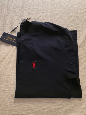 Svart t-shirt från Polo Ralph Lauren - Snygg svart t-shirt från Polo Ralph Lauren med klassisk röd logga broderad på bröstet. T-shirten har rund halsringning och korta ärmar. Tillverkad i mjuk bomull för en skön känsla och stilren look. Pris kan diskuteras 