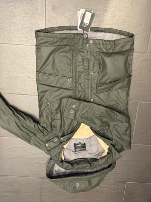 Pelle Petterson Levron Parka i färgen Dark Moss - Stilren grön vindjacka från PelleP med huva, dragkedja och tryckknappar. Jackan har flera praktiska fickor framtill och justerbara ärmslut. Perfekt för blåsiga dagar och outdoor-aktiviteter. Materialet är lätt och vattenavvisande. Sprillans ny jacka från PelleP. Nypris 4000kr, storlek S. Säljer den för 1500kr. Den har tags kvar. 