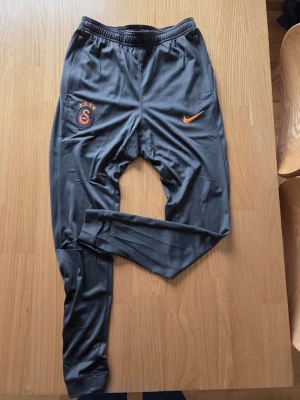 Nike gråa Galatasaray träningsbyxa, storlek S - Träningsbyxor från Nike i slim fit-modell. Byxorna har muddar vid bensluten och dragsko i midjan för justerbar passform. De är utrustade med dragkedjeförsedda fickor för säker förvaring av småsaker. Byxorna är prydda med Galatasaray-emblemet och Nike-logotypen.