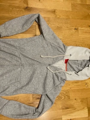 Björn Borg Ljusgrå zip hoodie med dragsko stl XSmall  - Snygg ljusgrå hoodie med dragkedja och huva med vita dragsnören. Insidan av huvan har ett rutigt mönster och en röd detalj vid halsen. Perfekt för en avslappnad och sportig stil. Mjuk bomullsblandning och två fickor framtill.