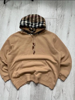 Beige burberry hoodie (Äkta) byten! - Snygg beige hoodie från Burberry med klassisk rutmönstrad huva i brunt, svart, vitt och rött. Hoodien har känguruficka framtill, ribbade muddar och dragsko i huvan. Perfekt för dig som vill ha en stilren och exklusiv look. Inga defekter 10/10 skick. Nypris 11000. Kan bytas mot hoodies/jackor i S/M