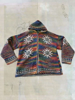 Flerfärgad stickad hoodie med solmönster - Unik stickad hoodie i regnbågsfärger med vita och svarta solmönster på både fram- och baksida. Tröjan har dragkedja, huva och ribbstickade muddar med mönsterdetaljer. Perfekt för dig som gillar färg och bohemisk stil.
