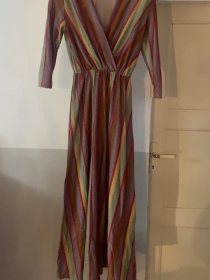 Randig långklänning från Adele Altman - Färgglad långklänning från Adele Altman med diagonala ränder i pastelliga toner av rosa, gult, grönt och beige. Klänningen har omlottliv, trekvartsärm och ett lätt glittrigt tyg som ger en extra touch. Perfekt för dig som gillar unika plagg med snygg passform.