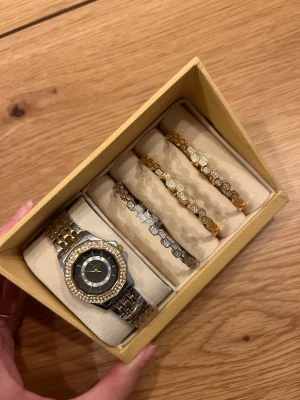 Klocka och armband set i guld- och silverfärg - Set med en tvåfärgad klocka och tre matchande armband. Klockan har ett kvartsurverk och är prydd med stenar runt boetten och på urtavlan. Armbanden har en bikakeliknande design med stenar, två i guldfärg och ett i silverfärg. Setet kommer i en presentförpackning. Setet är guldpläterat. 