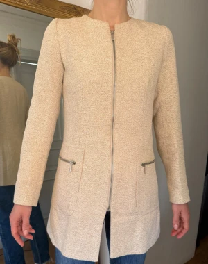 Beige kappa med dragkedja - Stilren kappa i beiget med lite guldig känsla. Två fickor med silverfärgade dragkedjor. Kappan har rundad halsringning och är tillverkad i ett strukturerat tyg som känns både mjukt och exklusivt.