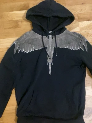 Svart hoodie Marcelo burlon - Snygg svart hoodie från Marcelo Burlon med unikt silverfärgat vingtryck över axlar och bröst. Klassisk huva med dragsko och känguruficka framtill. Perfekt för dig som vill sticka ut med en cool streetstyle-look.