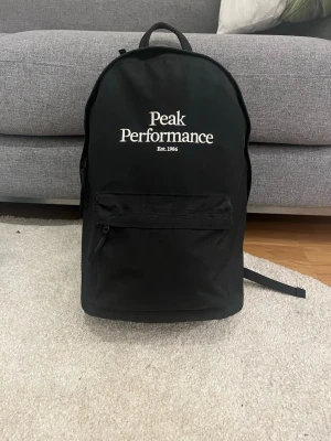 Svart ryggsäck Peak Performance - Snygg svart ryggsäck från Peak Performance med vit logga framtill. Klassisk form med ett stort huvudfack och en framficka med dragkedja. Justerbara, vadderade axelband och bärhandtag upptill. Tillverkad i slitstarkt tyg, perfekt för skolan eller stan.