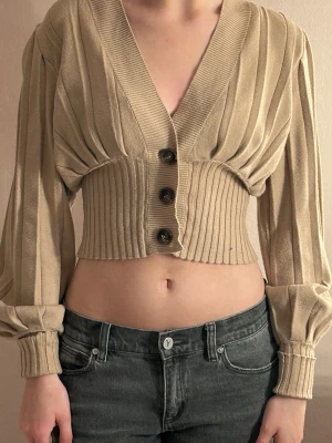 Beige cropped kofta från NA-KD - Trendig beige cropped kofta från NA-KD med bred ribbad midja, djup V-ringning och stora knappar framtill. Koftan har långa ärmar och är tillverkad i ett mjukt, stickat material. Perfekt för en snygg och avslappnad look.