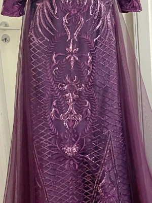 Lång lila klänning med brodyr - Elegant långklänning i mörklila med detaljerad brodyr och transparent tyll ovanpå. Klänningen har halvlånga ärmar och ett vackert mönster framtill som ger en lyxig känsla. Perfekt för dig som vill sticka ut med en unik och stilren look.