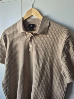 H&M pikétröja  - Säljer en beige ribbad pikétröja från H&M med klassisk krage och knappar framtill. Tröjan har korta ärmar och är tillverkad i mjuk bomull. Perfekt för en avslappnad men ändå stilren look.