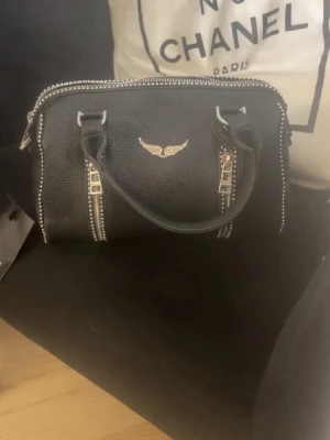 Svart handväska från zadig & voltaire - Cool svart handväska i skinnimitation med silvriga nitar längs dragkedjorna och ett vingemblem i metall framtill. Väskan har två handtag och detaljerade sömmar, perfekt för dig som gillar edgy stil. Från zadig & voltaire. 