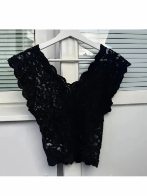 Svart spetsblus med v-ringning - Svart blus i spets med v-ringad hals och vågiga kanter. Ärmlös modell som sitter tajt och har transparent spetsmaterial. Perfekt att styla med jeans eller kjol för en trendig look.