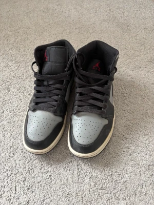Air Jordan 1 Mid svart/grå - Snygga Air Jordan 1 Mid sneakers i svart och grått läder med svarta skosnören och röd logga på sidan och plösen. Klassisk siluett med perforerad tåbox och vit sula. Perfekta för dig som gillar streetwear och ikoniska sneakers.