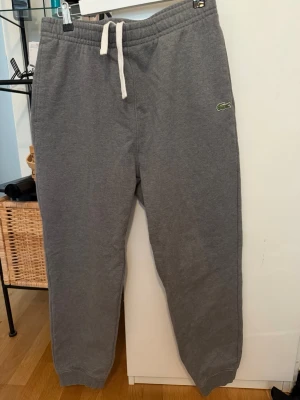 Grå mjukisbyxor från Lacoste - Snygga grå mjukisbyxor från Lacoste med klassisk krokodillogga på vänster ben. Byxorna har resår i midjan med vit dragsko, muddar vid bensluten och en bakficka. Perfekta för chill dagar och streetwear-stil.