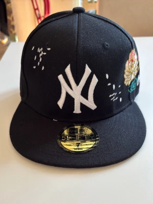 Svart New Era NY keps med brodyr - Snygg svart keps från New Era med stor vit NY-logga framtill och färgglad brodyr med blommor och moln på sidan. Flat peak och klassisk snapback-stil. Perfekt för dig som vill sticka ut med en unik accessoar.
