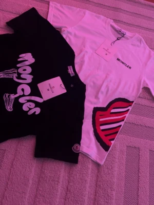 Moncler t-shirts med tryck - Två snygga t-shirts från Moncler, en svart med grafiskt tryck och logga på ärmen samt en vit med Moncler-tryck och stort rött och svart märke på sidan. Båda är kortärmade och i mjuk bomull, perfekta för en trendig streetwear-look.