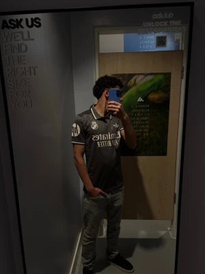 Svart Real Madrid t-shirt från Adidas - Snygg svart Real Madrid t-shirt från Adidas med vita detaljer och klubbmärke på bröstet. Tröjan har korta ärmar, klassisk krage och tryckt sponsorlogga framtill. Perfekt för dig som älskar fotboll och vill ha en sportig stil. Den är äkta, nyköpt med prislapp