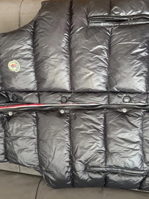 Svart dunväst från Moncler - Snygg svart dunväst från Moncler med klassisk quiltad design och emblem på bröstet. Västen har dragkedja, tryckknappar och innerficka med röd dragkedja. Fodrad med dun för extra värme och har en cool serietidningsdetalj på insidan.