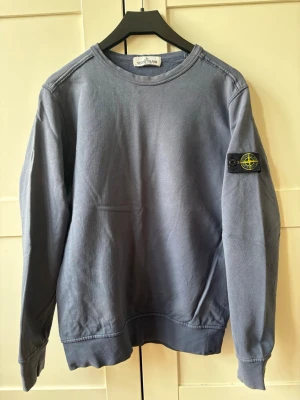 Blå sweatshirt från Stone Island - Snygg blå sweatshirt från Stone Island med klassisk logotyp-patch på vänster ärm. Tröjan har rund halsringning, ribbade muddar och är tillverkad i mjukt bomullsmaterial. 