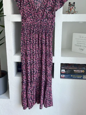 Blommig långklänning från Pernilla Wahlgren - Långklänning med blommigt mönster i rosa och blått från Pernilla Wahlgren. Klänningen har v-ringning, korta ärmar och smockad midja för en bekväm passform. Perfekt för sommarens alla tillfällen.