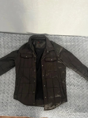 Olivgrön pufferjacka från Dressmann - Olivgrön pufferjacka från Dressmann med rutmönstrad quiltning, två bröstfickor med lock och knappar samt två sidofickor. Jackan har hög krage och stängs med tryckknappar. Perfekt för kalla vinterdagar.