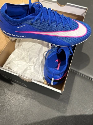 Nike mercurial fotbollsskor - Snygga Nike mercurials fotbollsskor i blått med vit och rosa swoosh. Skorna har en strömlinjeformad design, snörning och ovandel i syntetmaterial för bra passform och grepp på planen. Perfekta för dig som vill sticka ut på matchen. Helt nya oanvända , nypris 2899, mitt pris 1800 kr.