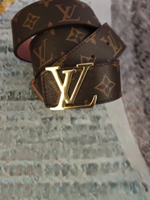 L V brunt monogram bälte - Snyggt bälte från med klassiskt brunt monogrammönster och guldigt L V-spänne. Baksidan är ljusrosa och bältet är gjort i skinn. Perfekt accessoar för att lyfta din outfit med en lyxig touch.