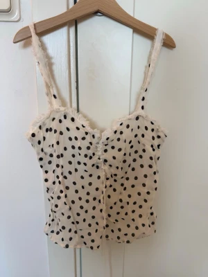 Prickig linnetopp från Zara - Söt linnetopp i vit med svarta prickar och tunna axelband. Toppen har spetsdetaljer längs kanterna och en lätt figurnära passform. Perfekt för en trendig och avslappnad look.