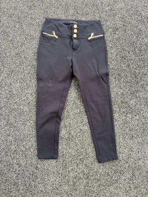Svarta leggings med guldknappar - Snygga svarta leggings med hög midja och tre guldknappar framtill. Två dekorativa guldfärgade dragkedjor vid fickorna och en bakficka med gulddetaljer. Stretchigt material som sitter tajt och bekvämt. Passar till 155 cm långa 