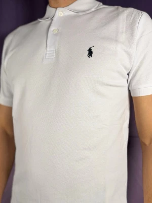Vit pikétröja från Ralph Lauren - Klassisk vit pikétröja från Ralph Lauren med korta ärmar och två knappar vid kragen. Tröjan har den ikoniska broderade loggan i svart på bröstet och är tillverkad i ett svalt pikématerial som passar perfekt till sommaren.