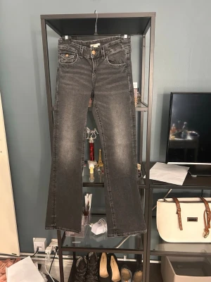 Grå bootcut jeans med broderi - Snygga grå bootcut jeans med slitningar och broderade detaljer på bakfickorna. Jeansen har klassisk femficksdesign, knappgylf och kontrastsömmar. Perfekta för dig som gillar en avslappnad men trendig stil.