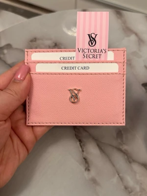Rosa korthållare från Victoria's Secret - Supersnygg rosa korthållare från Victoria's Secret i ett stilrent och smidigt format. Har tre fack för kort och ett elegant VS-metallogo framtill. Materialet är strukturerat konstläder som känns lyxigt och trendigt.
