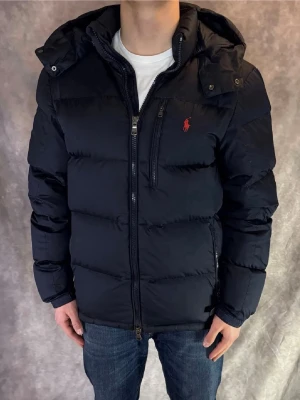 Ralph Lauren Jacka - Säljer en stilren svart dunjacka från Polo Ralph Lauren. Jackan har en klassisk och tidlös design med röd broderad logga på bröstet, avtagbar huva och flera praktiska fickor med dragkedja. Perfekt för höst och vinter då den är både varm och bekväm samtidigt som den sitter snyggt på kroppen.