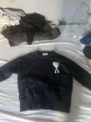 Mörkblå Ami Paris tröja med hjärtdetalj - Snygg mörkblå tröja från Ami Paris med ett vitt hjärta och bokstaven A broderat på bröstet. Tröjan har rund halsringning, långa ärmar och breda ribbade muddar vid ärmslut och nederkant. Perfekt för en stilren och trendig look.