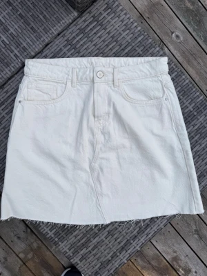 Gräddvit jeanskjol från VILA - Snygg gräddvit jeanskjol från VILA med klassisk femficksdesign och rå kant nertill. Kjolen har knapp och dragkedja framtill samt bälteshällor i midjan. Perfekt för dig som gillar en enkel och stilren look. Helt ny!