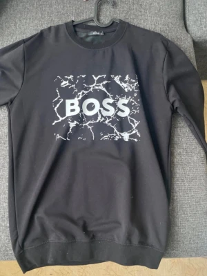 Svart sweatshirt från BOSS - Svart sweatshirt från BOSS med vit marmorerad print och stor logga framtill. Tröjan har rund halsringning, långa ärmar och ribbade muddar vid ärmslut och nederkant. Perfekt för en cool och avslappnad stil. Står att den är XL men tycker den är mer L eller M