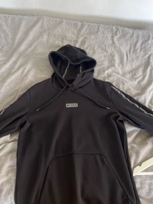 Svart hoodie från HUGO med logga - Svart hoodie från Hugo med logga på bröstet och längs ärmarna. Klassisk huva med dragsko och stor magficka framtill. Skön och stilren modell som passar perfekt till streetwear. Tillverkad i mjukt material för maximal komfort.