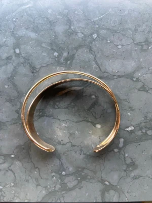 Guldigt öppet armband i metall - Snyggt armband i guldig metall med öppen design och två parallella band som ger en modern och stilren look. Perfekt accessoar för att lyfta din outfit med en touch av elegans. Armbandet är stelt och formas runt handleden.