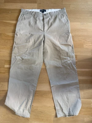 Beige chinos från Polo Ralph Lauren - Snygga beige chinos från Polo Ralph Lauren i klassisk rak modell. Tillverkade i mjuk bomull med knappgylf och bälteshällor. Två sidofickor fram och två bakfickor med knapp. Perfekta för en clean och avslappnad stil.  Lite nötta nere vid benslutet se bild. Storlek 36/30