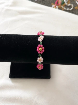 Blommigt pärlarmband i rosa och guld -  armband med blommor av pärlor i rosa, ljusrosa och grönt. Armbandet har guldfärgade detaljer och justerbart kedjelås. 