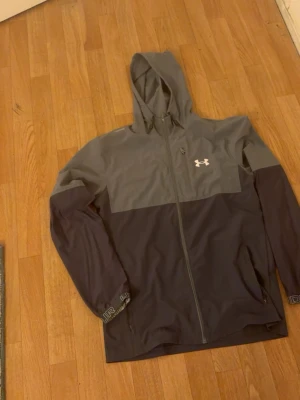 Grå vindjacka från Under Armour - Snygg tvåfärgad vindjacka från Under Armour i mörkgrått och ljusgrått. Jackan har huva, hel dragkedja framtill och logga på bröstet. Perfekt för blåsiga dagar och sportiga outfits. Materialet är lätt och vattenavvisande.