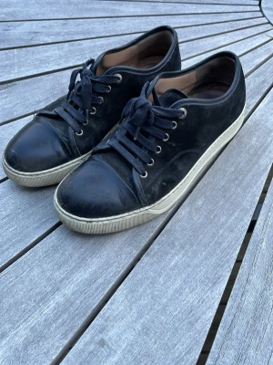 Svarta sneakers med mockadetaljer - Okej kvalite Lanvins med mocka och tåparti i slätt läder. Skorna har mörkblåa skosnören, vita sulor och diskreta sömdetaljer. Perfekta för dig som gillar en ”beat up” sko stil ( lcke glansig tåparti )